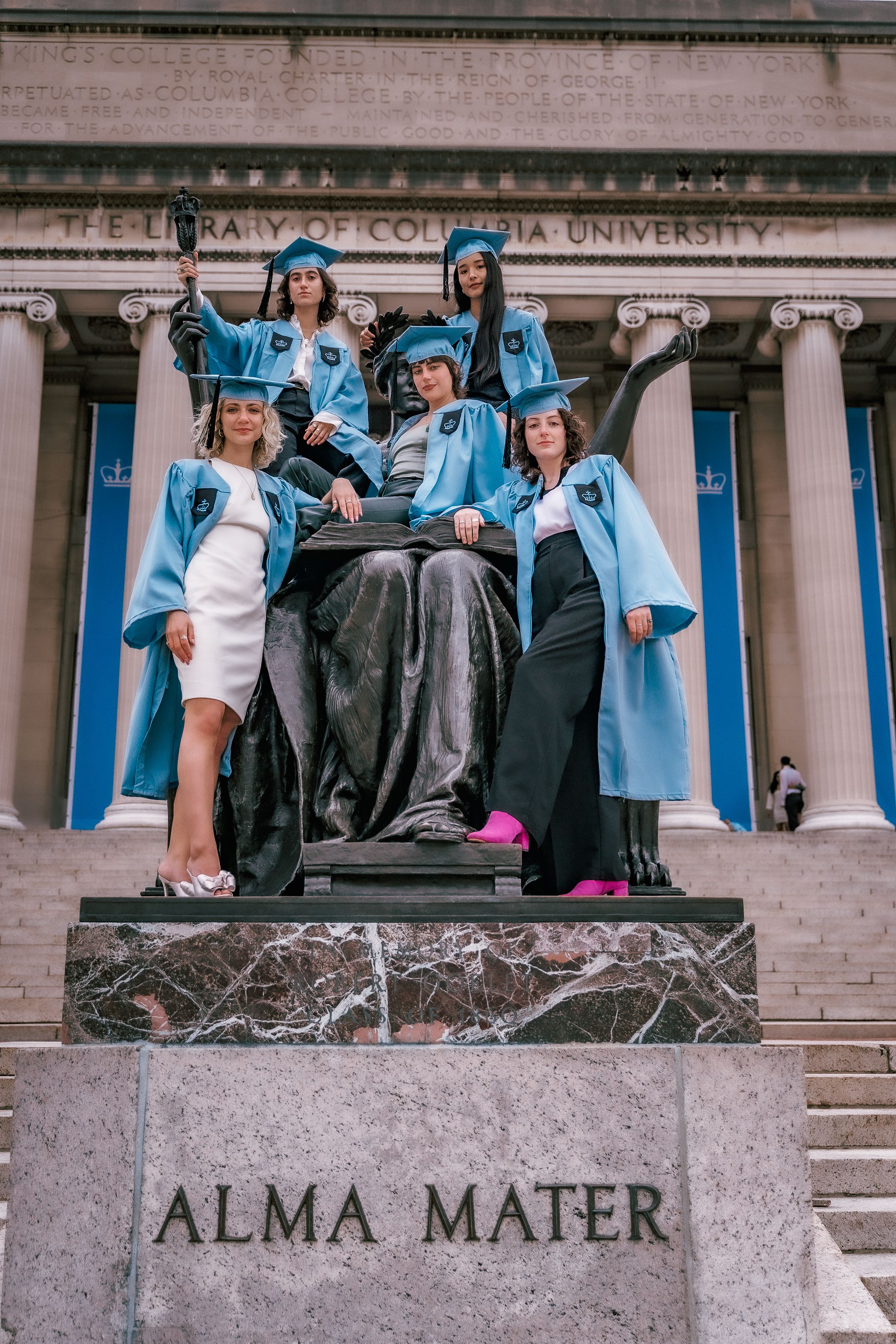 Columbia University graduates pose on the iconic Alma Mater statue wearing light blue graduation gowns.