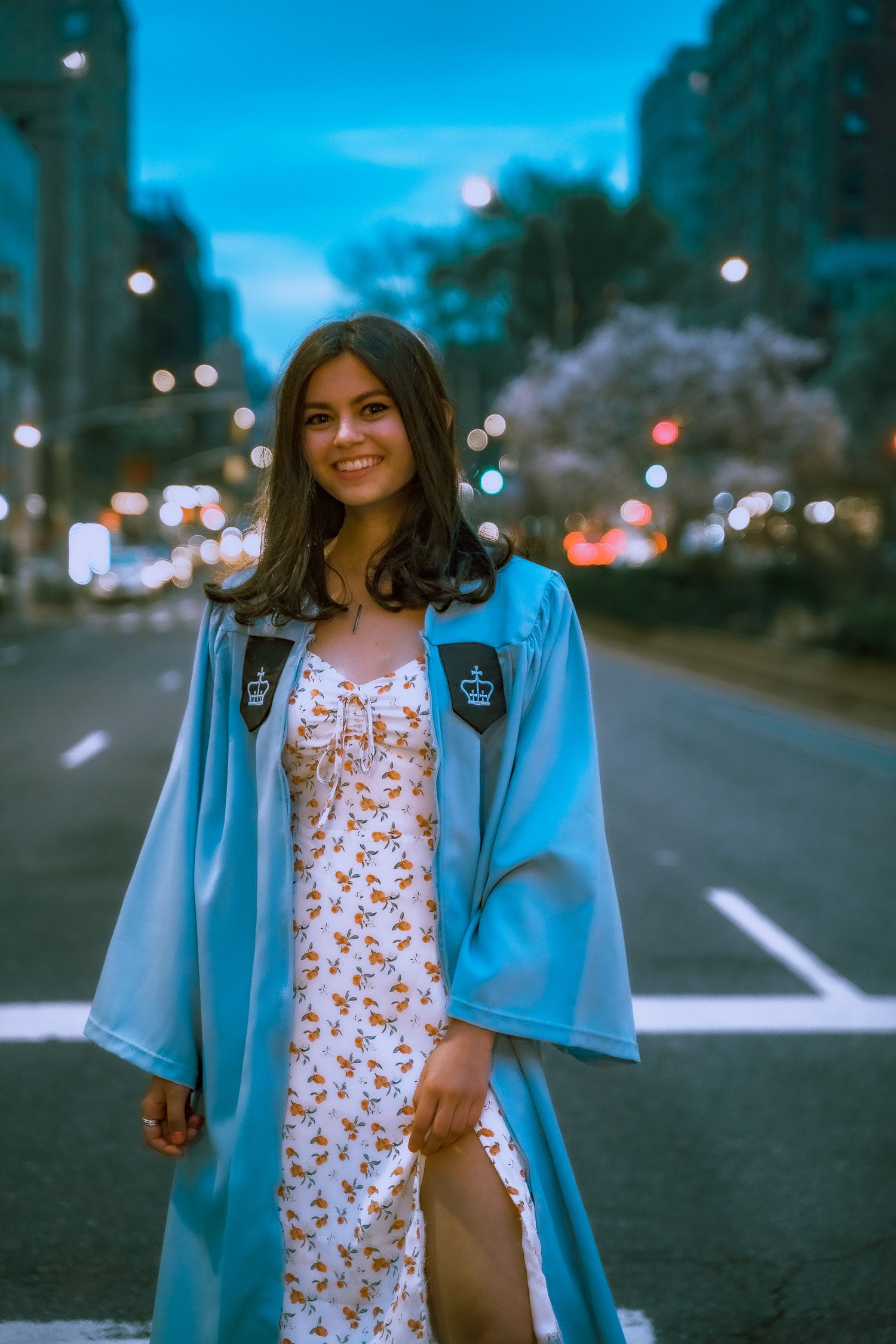 Graduate in light blue gown and floral dress poses on city street at dusk.