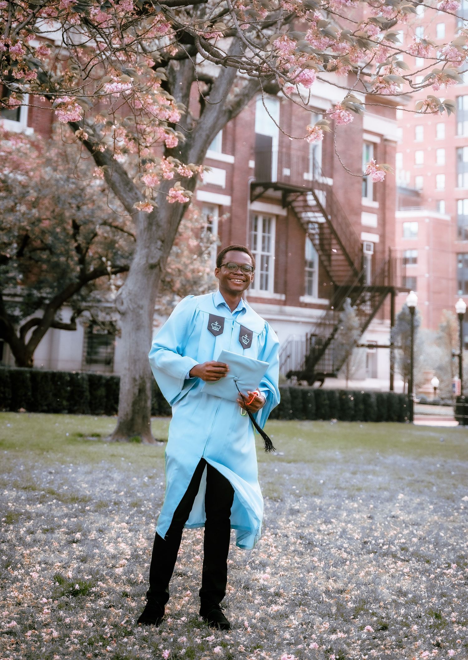 Graduate in light blue cap and gown smiles while holding diploma under blooming cherry trees.