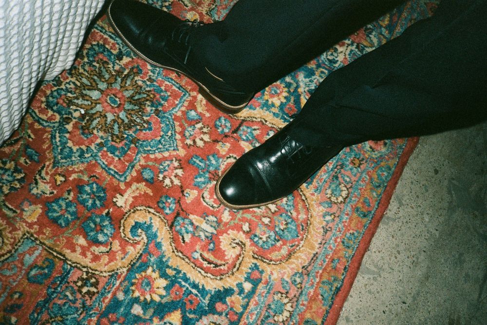 A black leather dress shoe rests on an ornate vintage rug with red and turquoise floral patterns.