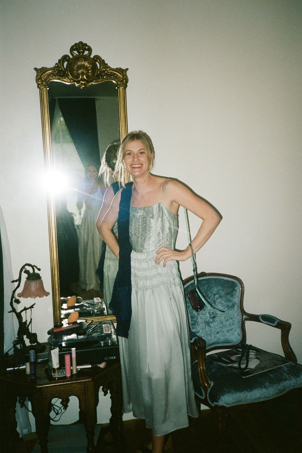 A formal dress reflected in an ornate gold-framed mirror next to a blue velvet chair.