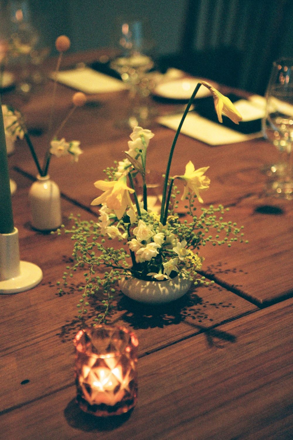 Small vases with yellow daffodils and greenery arranged on a wooden table with candlelight.