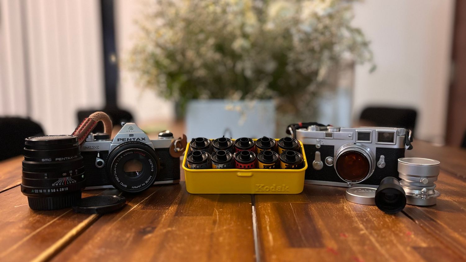 Two vintage cameras and a shot glass arranged on a wooden table with a yellow bowl of berries and flowers in the background.