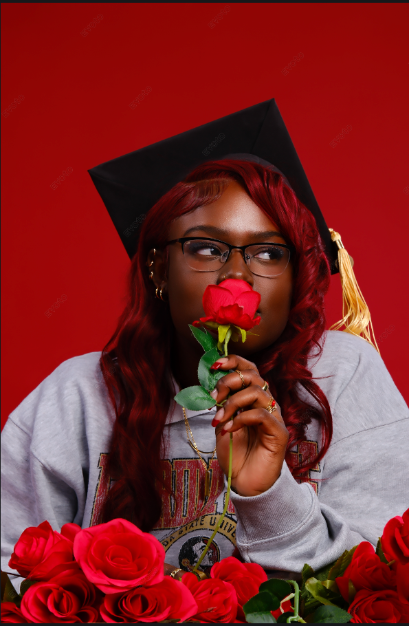 Graduate wearing a gray sweatshirt smells a red rose against a deep red background with roses in the foreground.