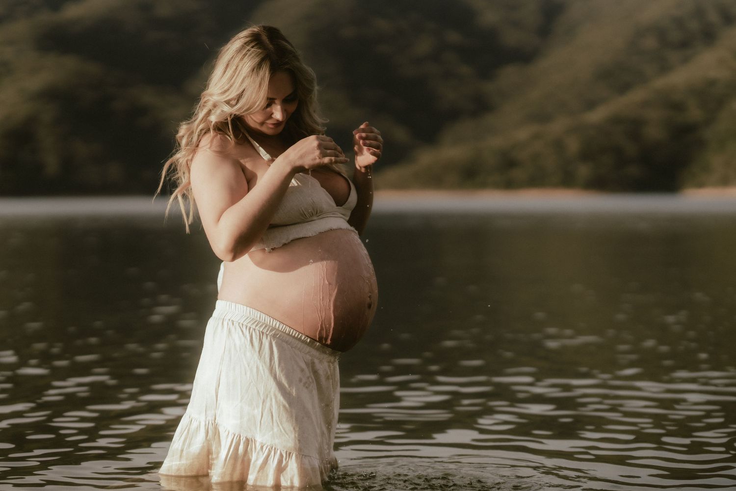 Dreamy maternity portrait by lake wearing white dress in golden light.