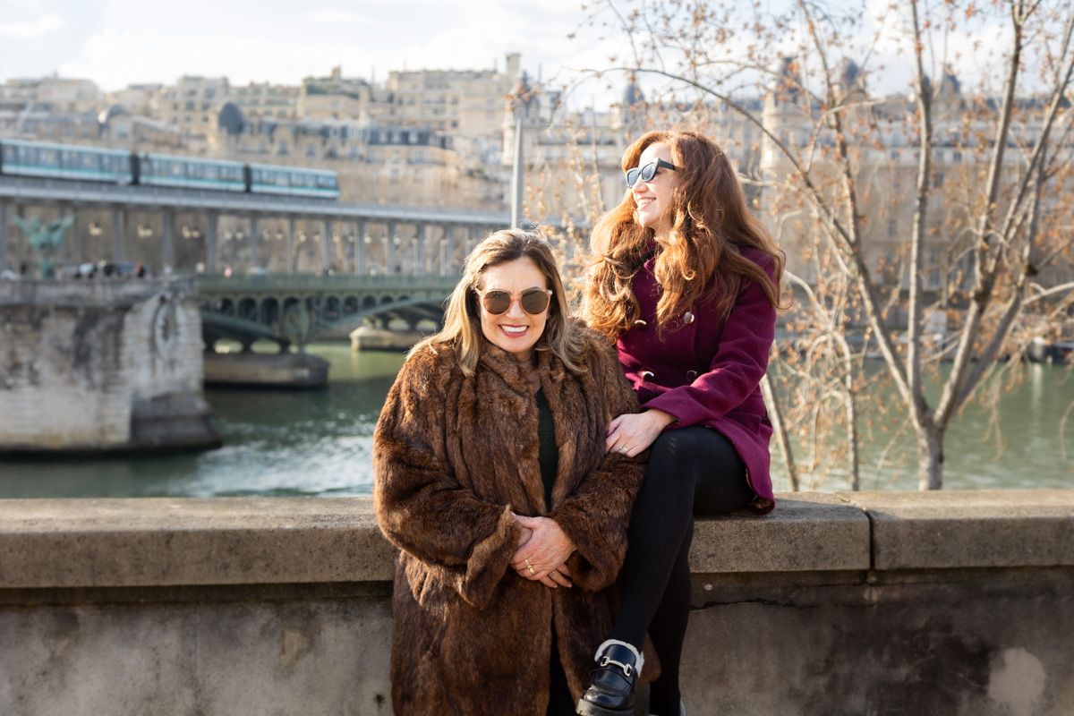 Two friends enjoying a sunny winter day along the Seine River in Paris with a stone bridge in the background.