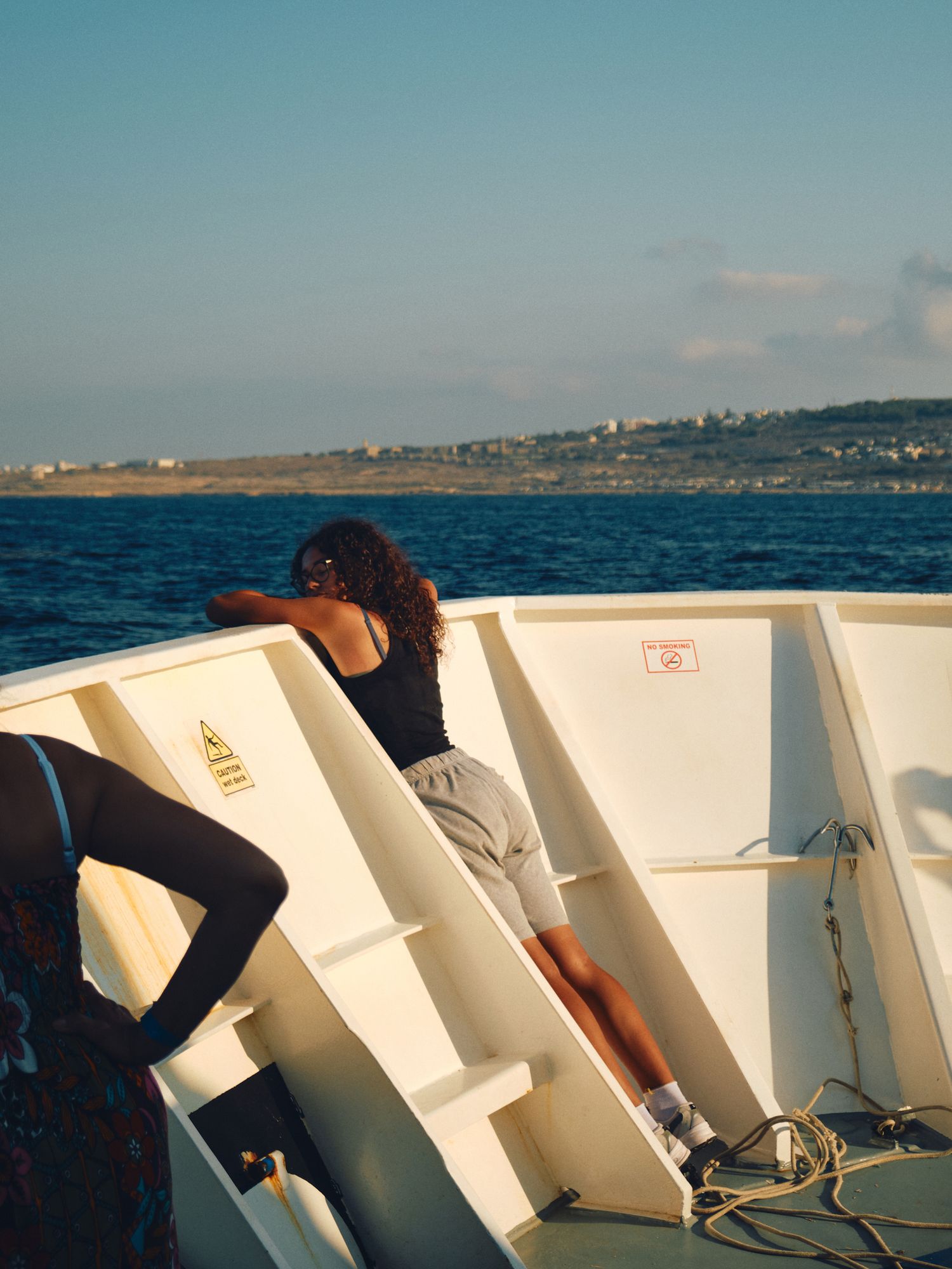 Someone sits on a boat deck with white seats while sailing across blue ocean waters near a coastal shoreline on a clear day.