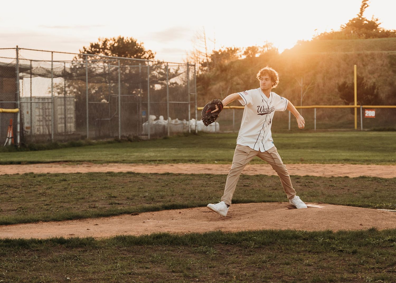 Baseball player winding up for a pitch on a dirt mound during golden hour sunset at a local baseball field.