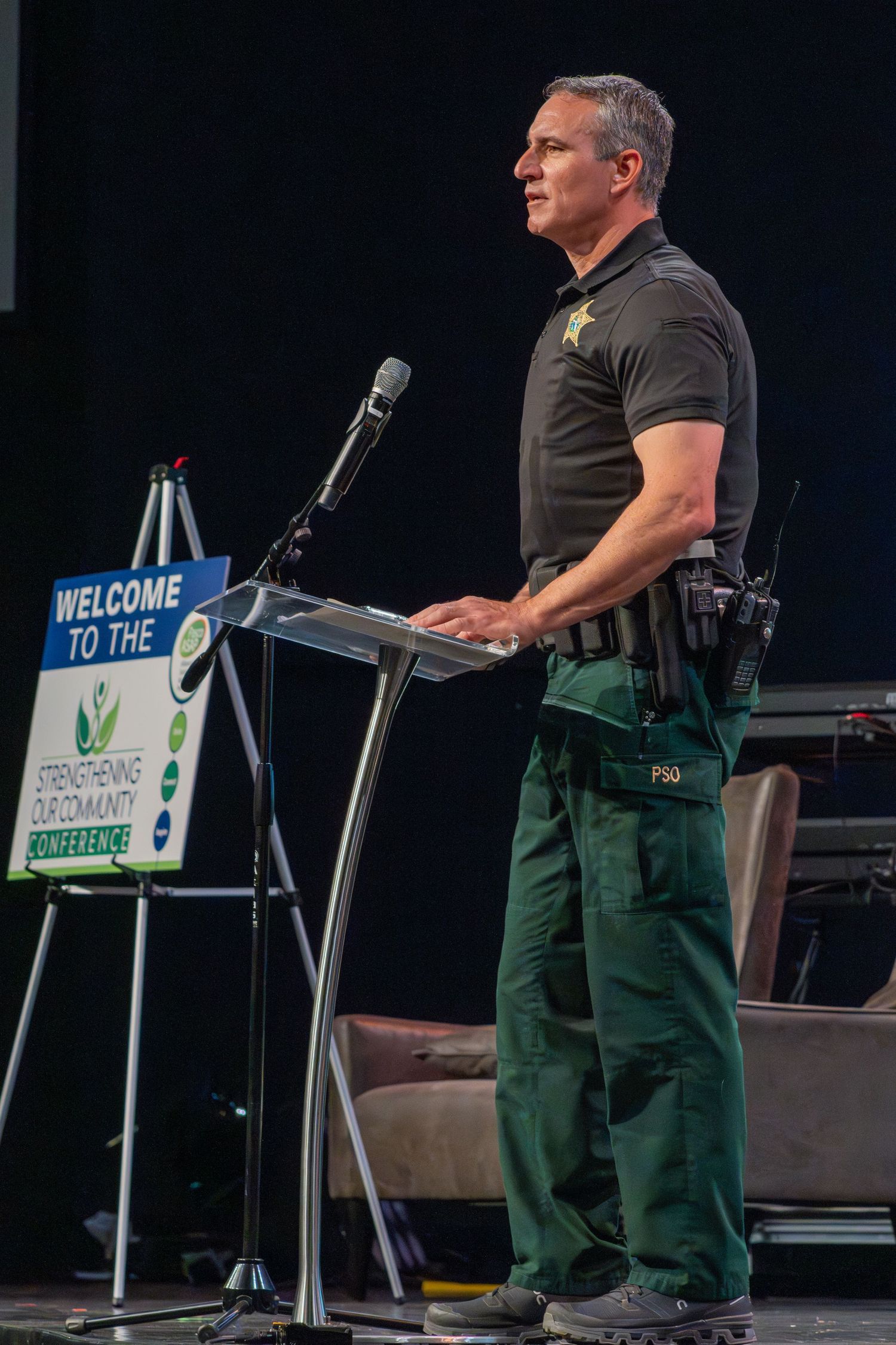 Speaker presenting on stage with green signage and projection screens in dark auditorium setting.