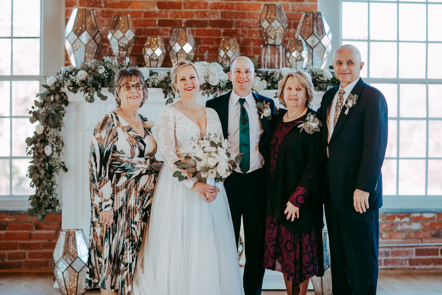 Formal wedding group photo with exposed brick wall and floral arch backdrop.