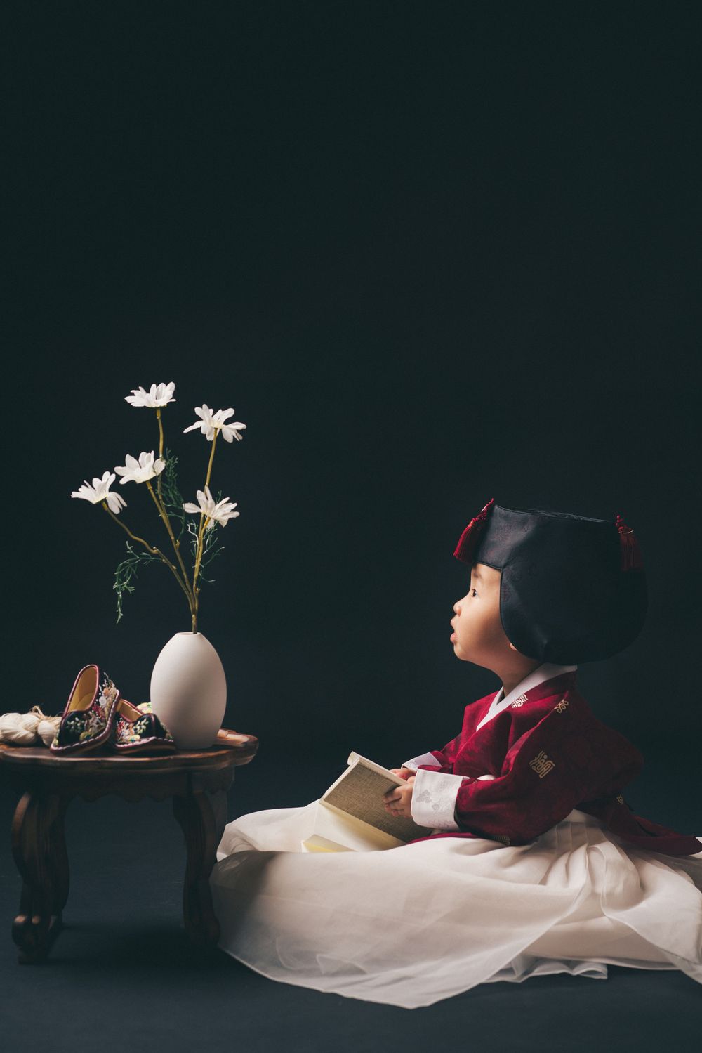 Child in traditional Korean hanbok dress sitting beside a small table with white daisies in a vase.
