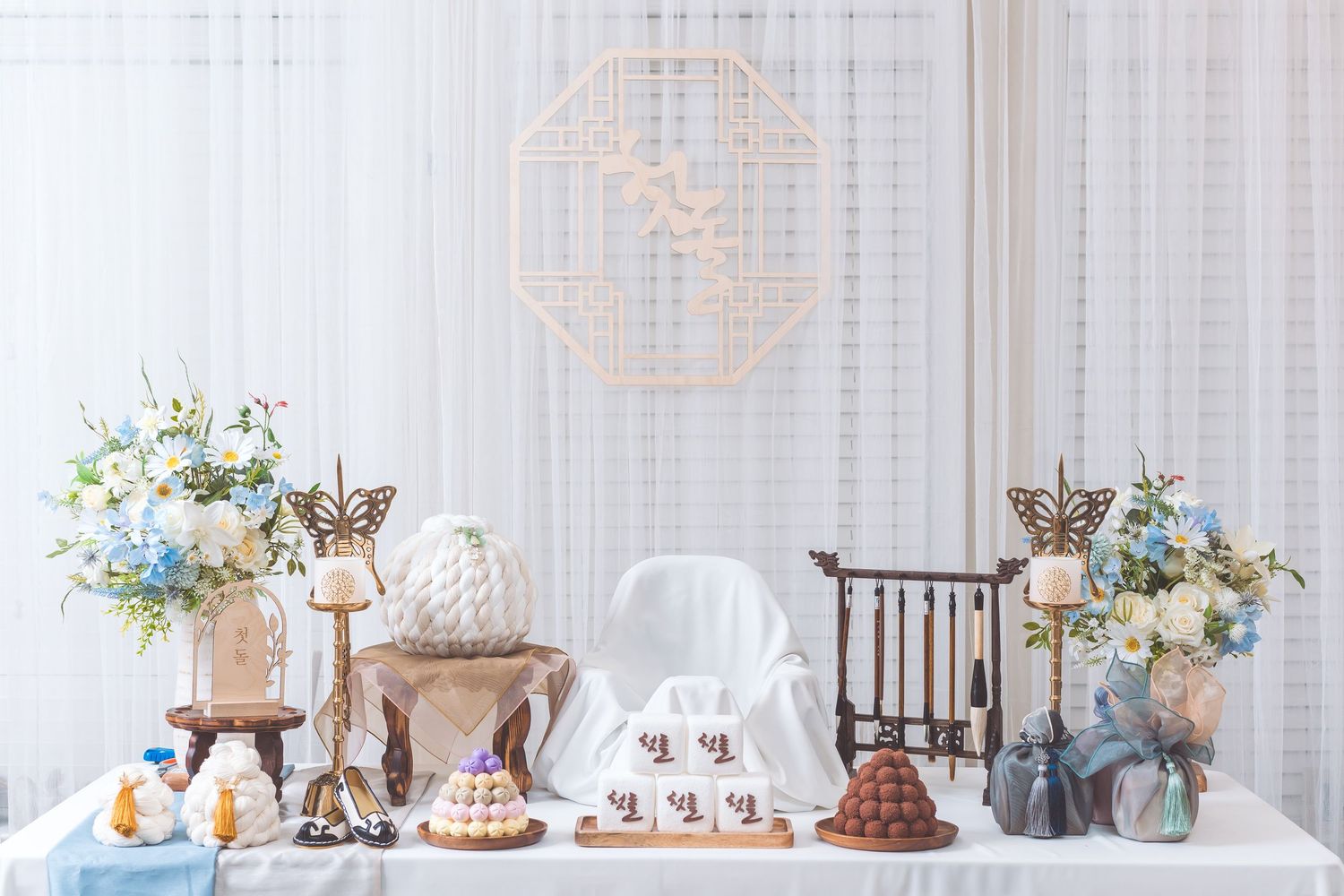 Dessert table setup with floral arrangements, tiered cakes, and white tablecloth against sheer curtains.