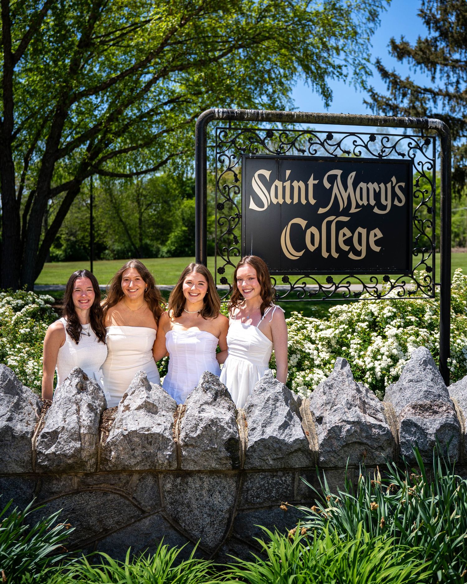 A group in white and blue dresses sits together on a stone wall at Saint Mary's College surrounded by spring greenery.