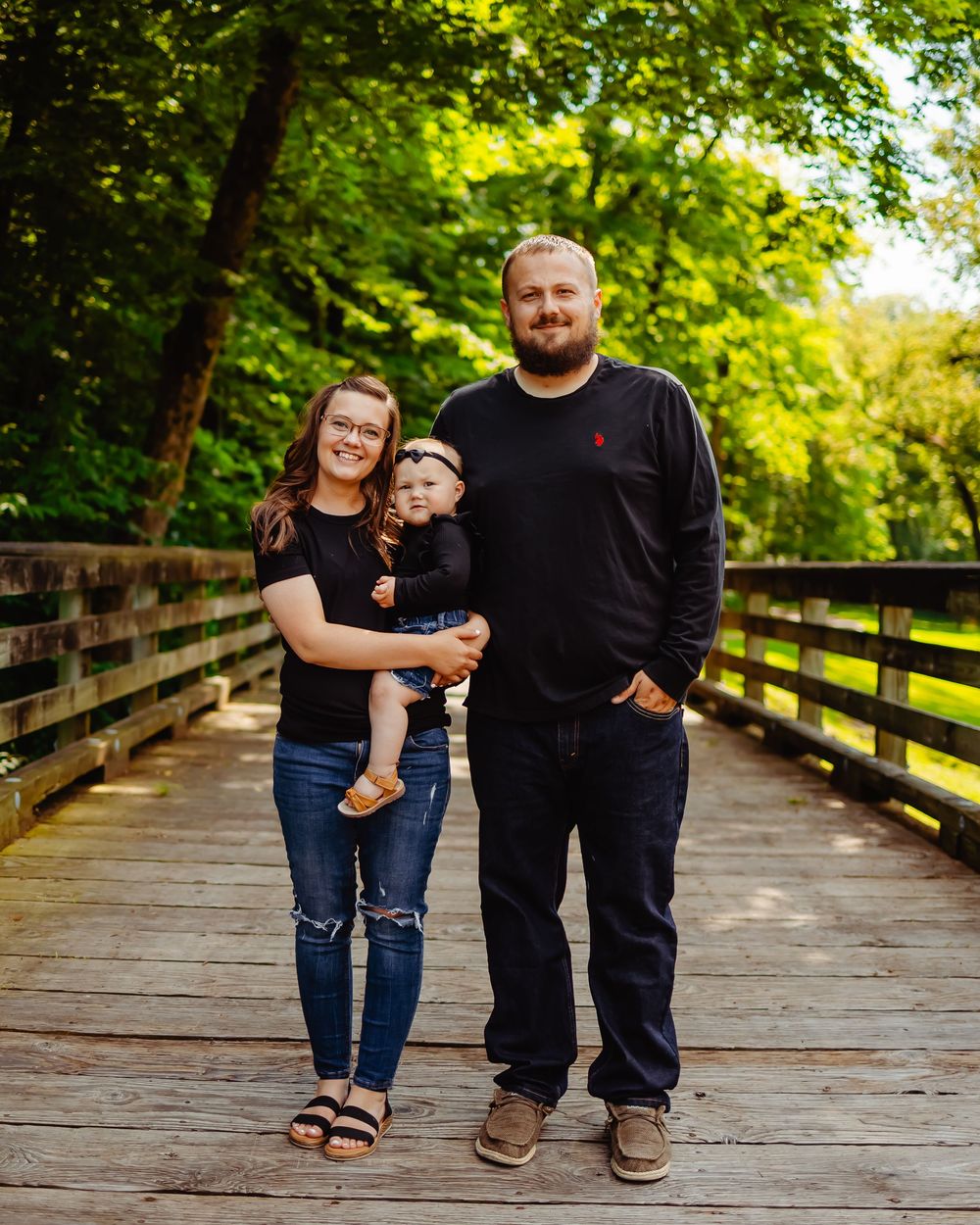 A family stands together on a wooden bridge walkway surrounded by lush green trees in a park setting.