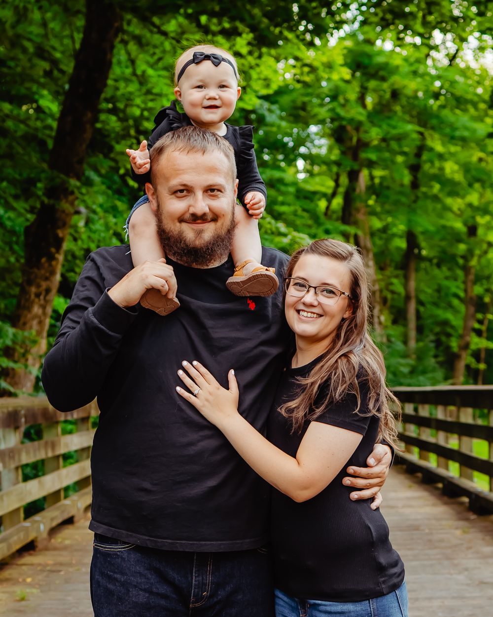A family poses together on a wooden bridge surrounded by lush green forest on a bright sunny day.