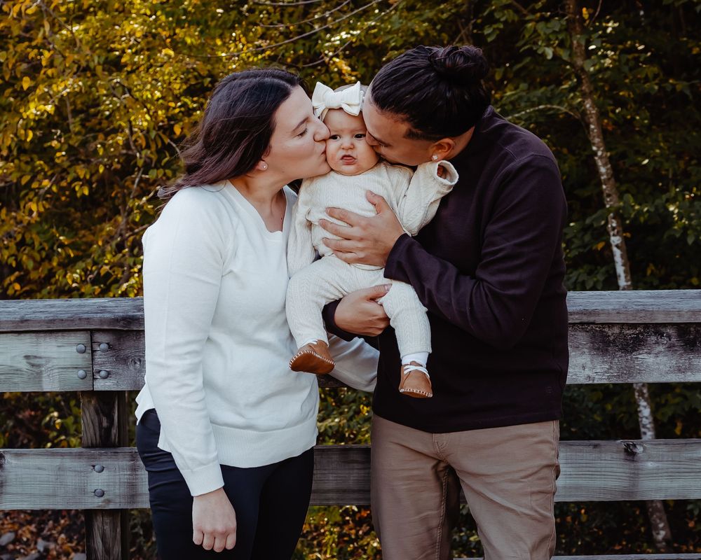 A couple in autumn shares tender moments with their baby while sitting on a wooden bench in a park.