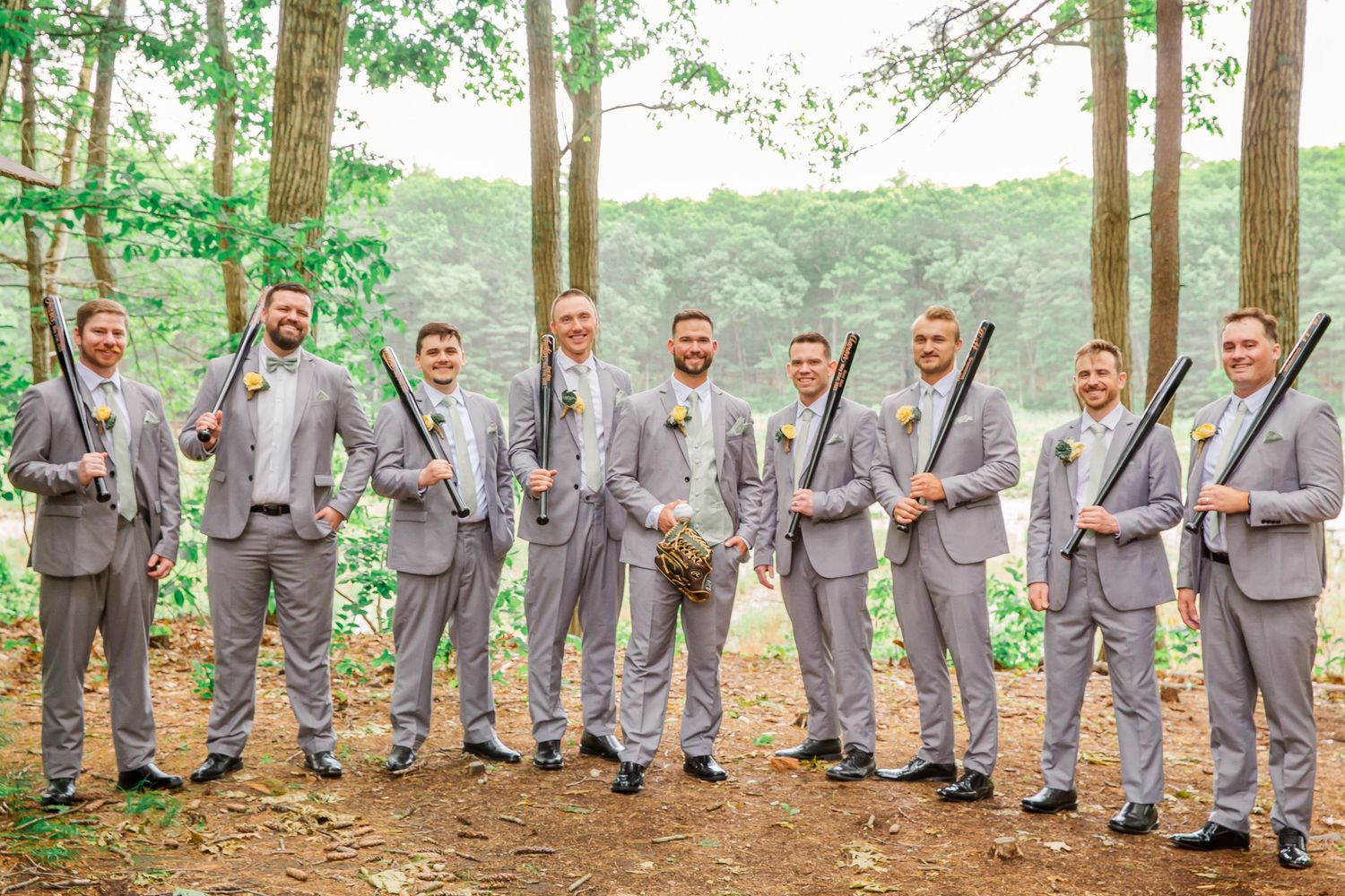 Nine groomsmen wearing matching light gray suits and ties pose together in a wooded outdoor setting.
