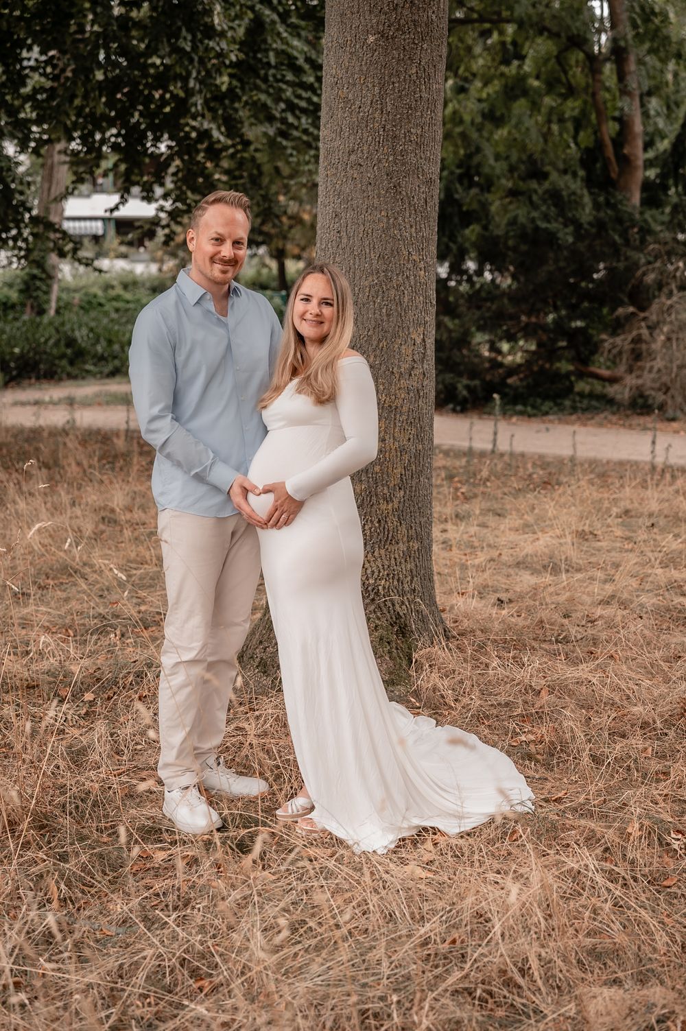 A couple poses together against a tree trunk, with the woman wearing a flowing white maternity gown in an outdoor setting.