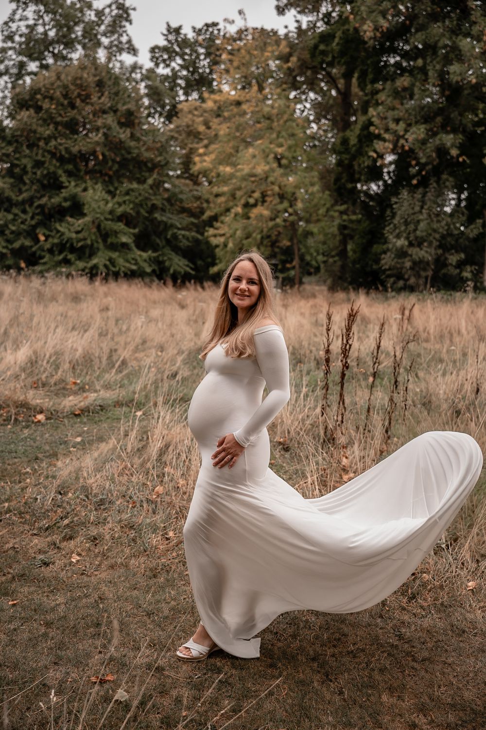 A woman in a flowing white dress poses gracefully in a rustic field with autumn foliage in the background.