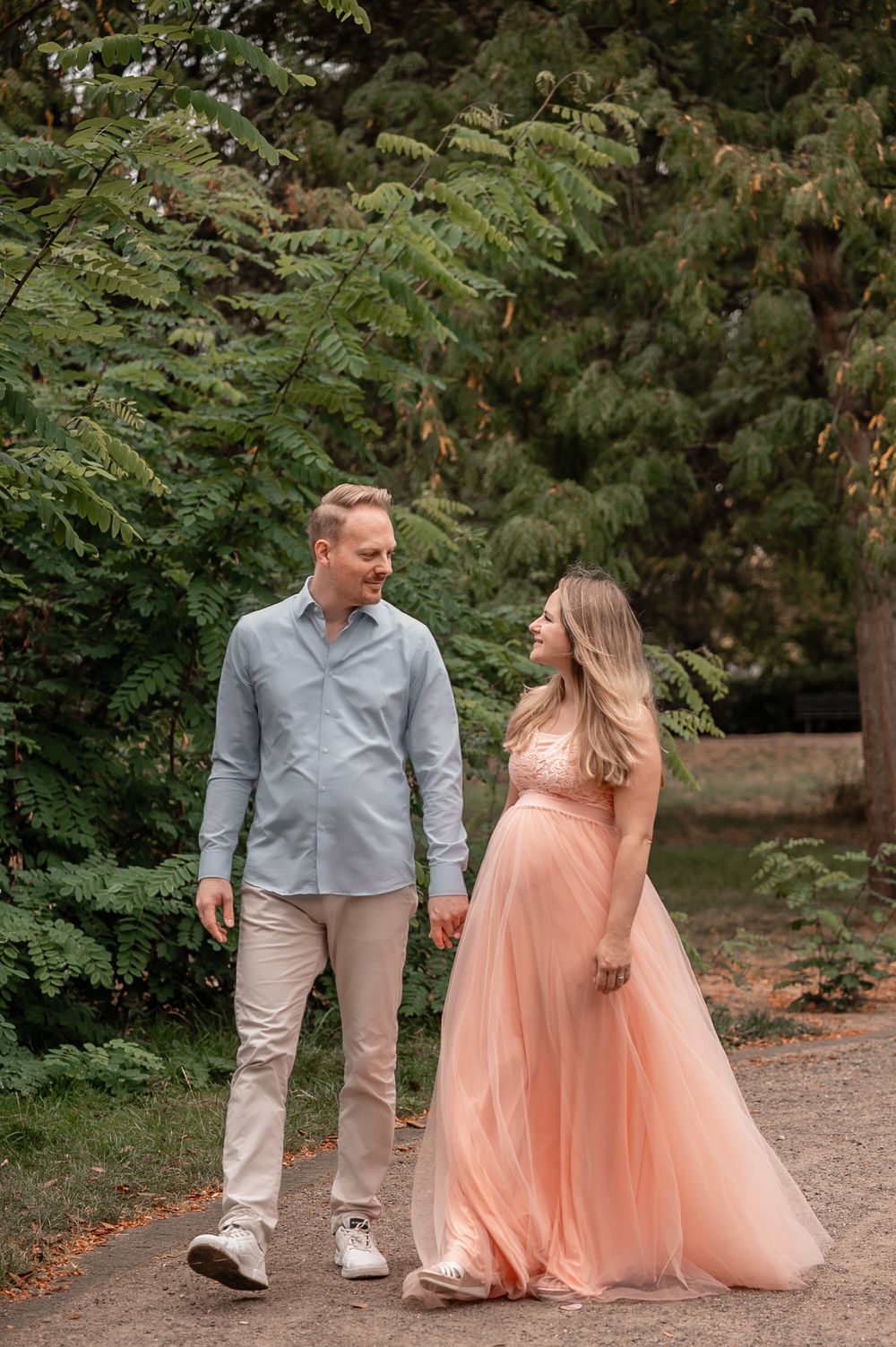 A couple in formal attire walks through a garden path, with the person in a peach-colored flowing dress.