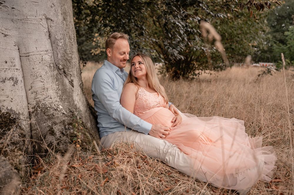 A couple in casual attire poses for romantic maternity photos in a rustic outdoor setting with tall grass.