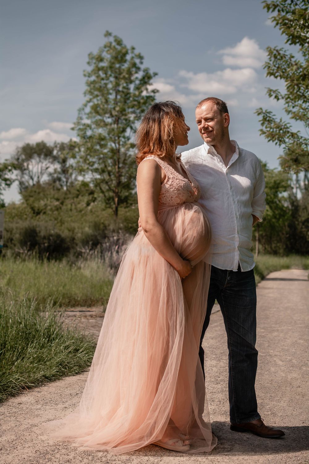 A couple shares an intimate moment on a path, with the expectant mother wearing a flowing peach gown.