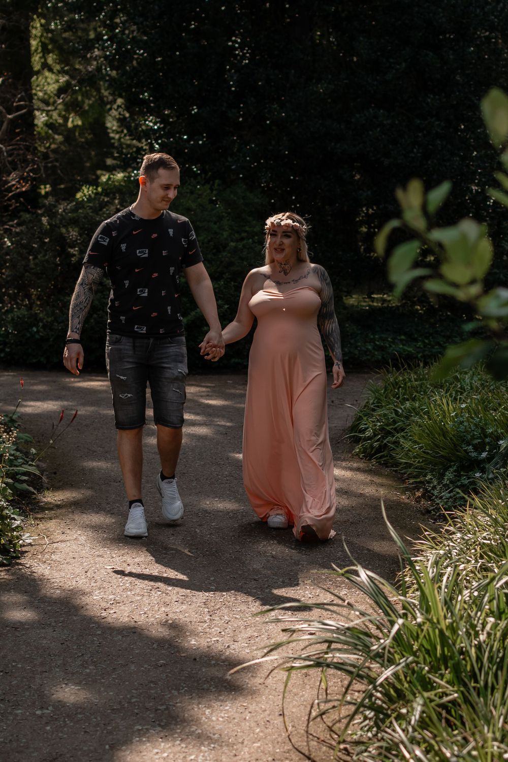 A couple walks hand in hand along a sunlit garden path during a maternity photoshoot.