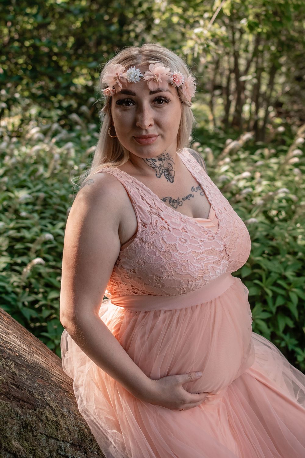 An expectant mother in a pink lace dress poses in a woodland setting.