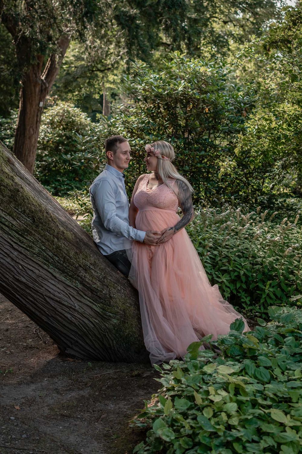 A romantic maternity photo of a couple posed against a fallen tree in a lush forest setting.