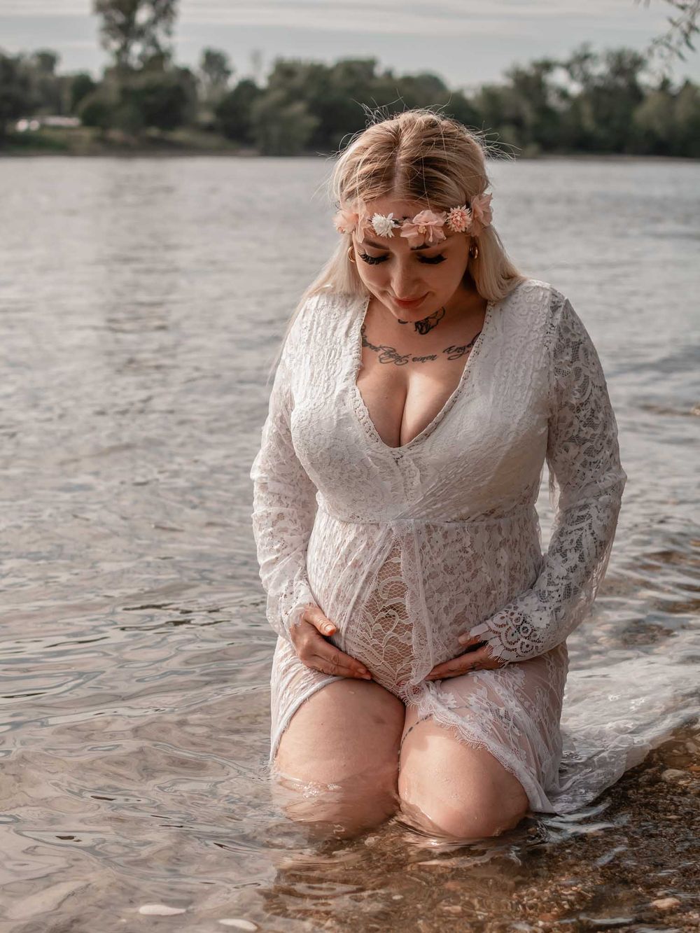 A person in a white lace dress kneels in shallow water during a romantic outdoor photoshoot.
