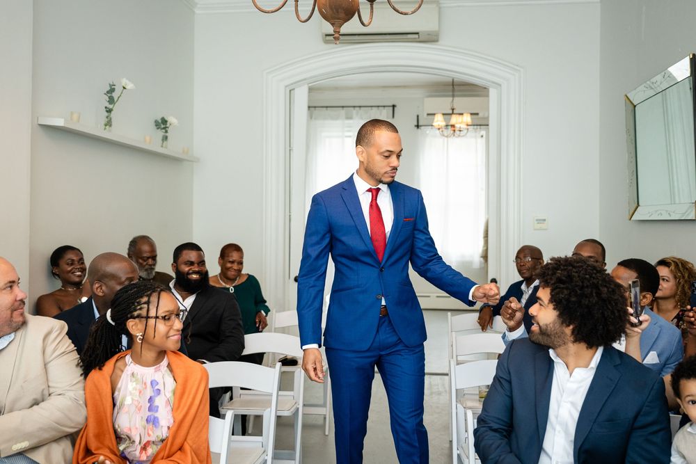 People gather in a bright white room as a man in a vibrant blue suit stands and gestures during an event.