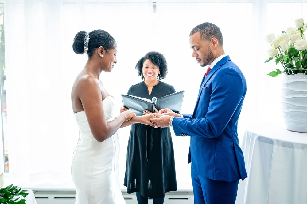 A couple stands at a wedding ceremony with an officiant in black robes as they exchange rings during their vows.