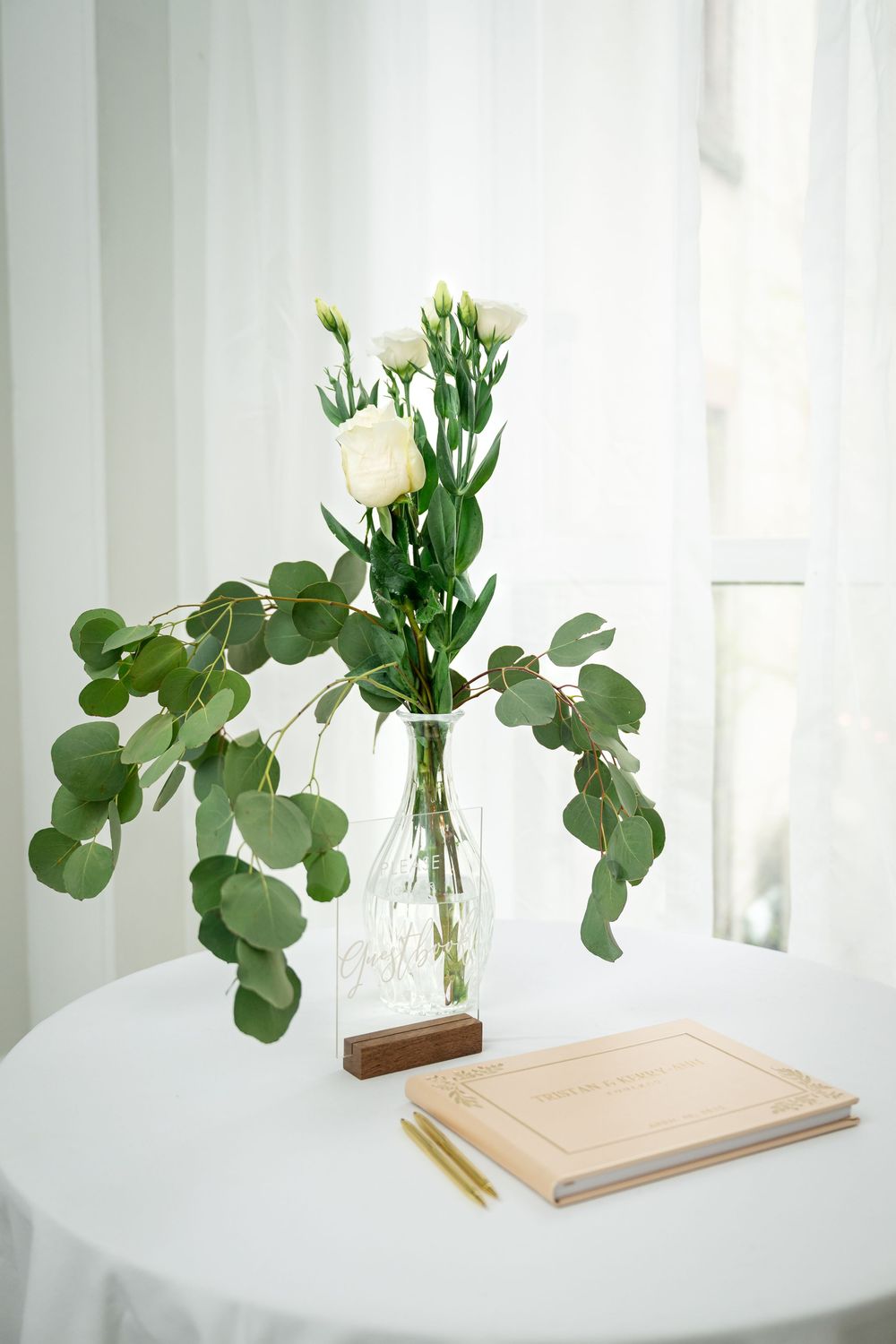 A minimalist vase with white flowers and eucalyptus leaves sits on a white table next to a notebook and pencil.