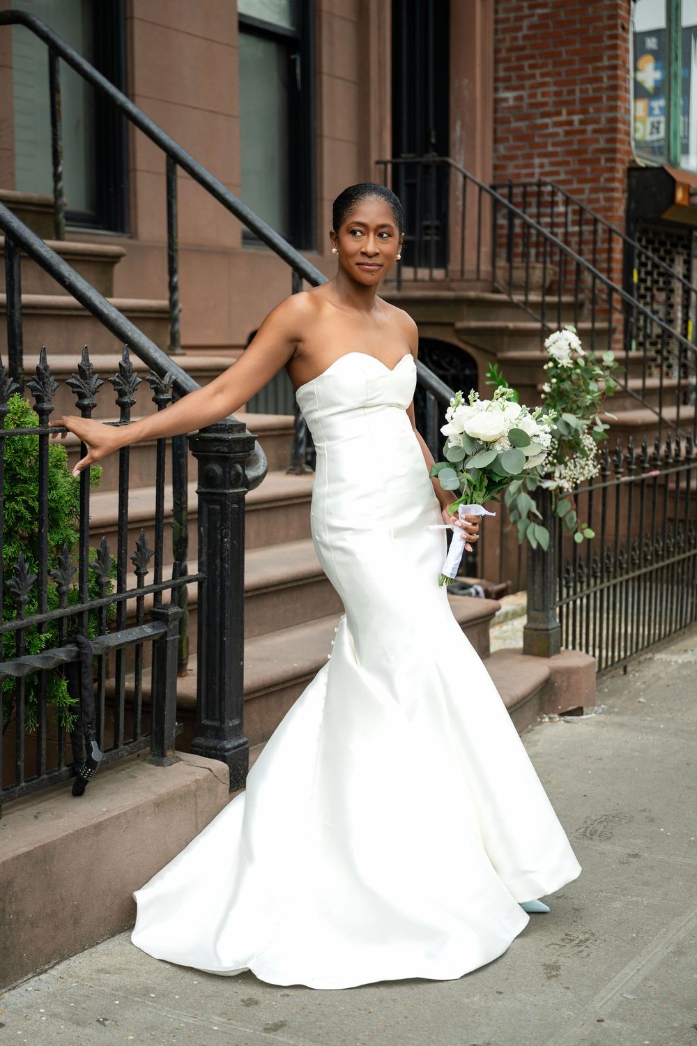 A woman in an elegant white strapless mermaid wedding gown holds a bouquet while standing on brownstone steps.