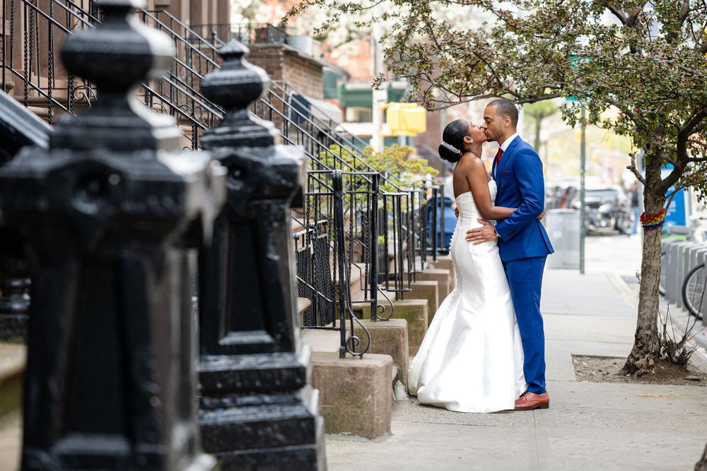 A wedding couple embraces on a city sidewalk with classic brownstone architecture in the background.