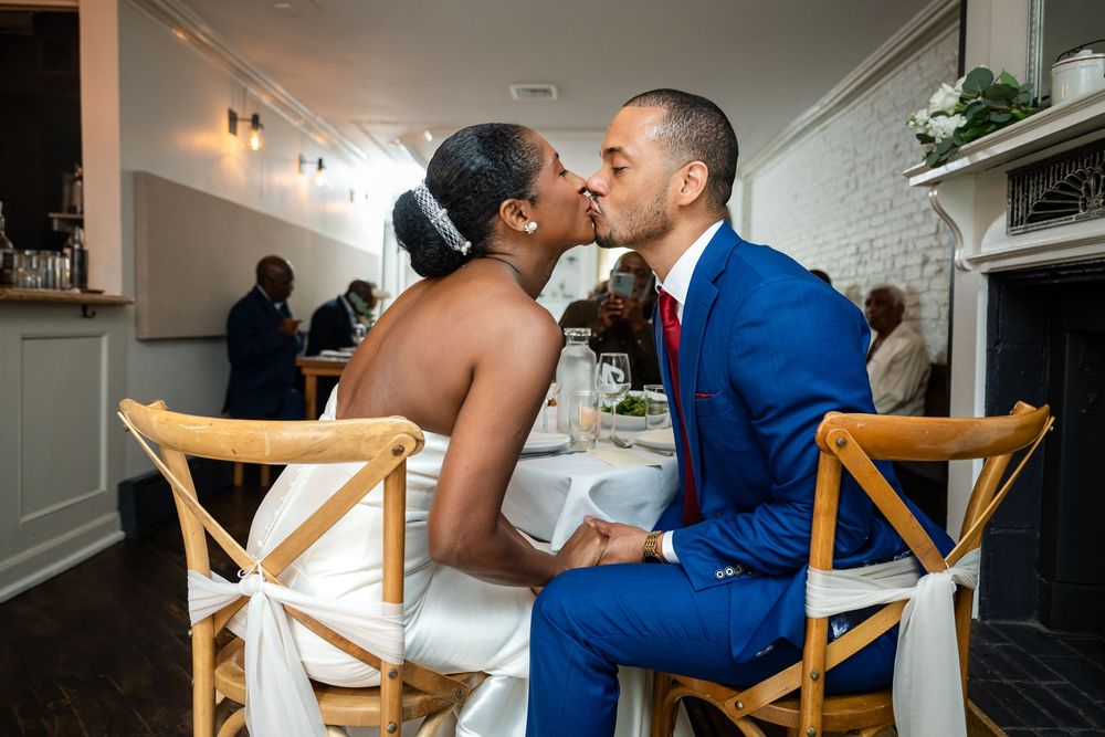 Newlywed couple sharing intimate kiss while seated at wedding reception in elegant venue.