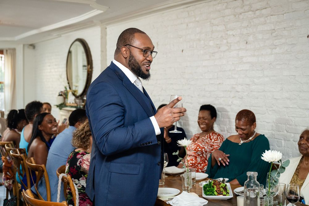 A speaker in a navy suit gestures animatedly during a celebratory dinner gathering at a bright restaurant with white walls.