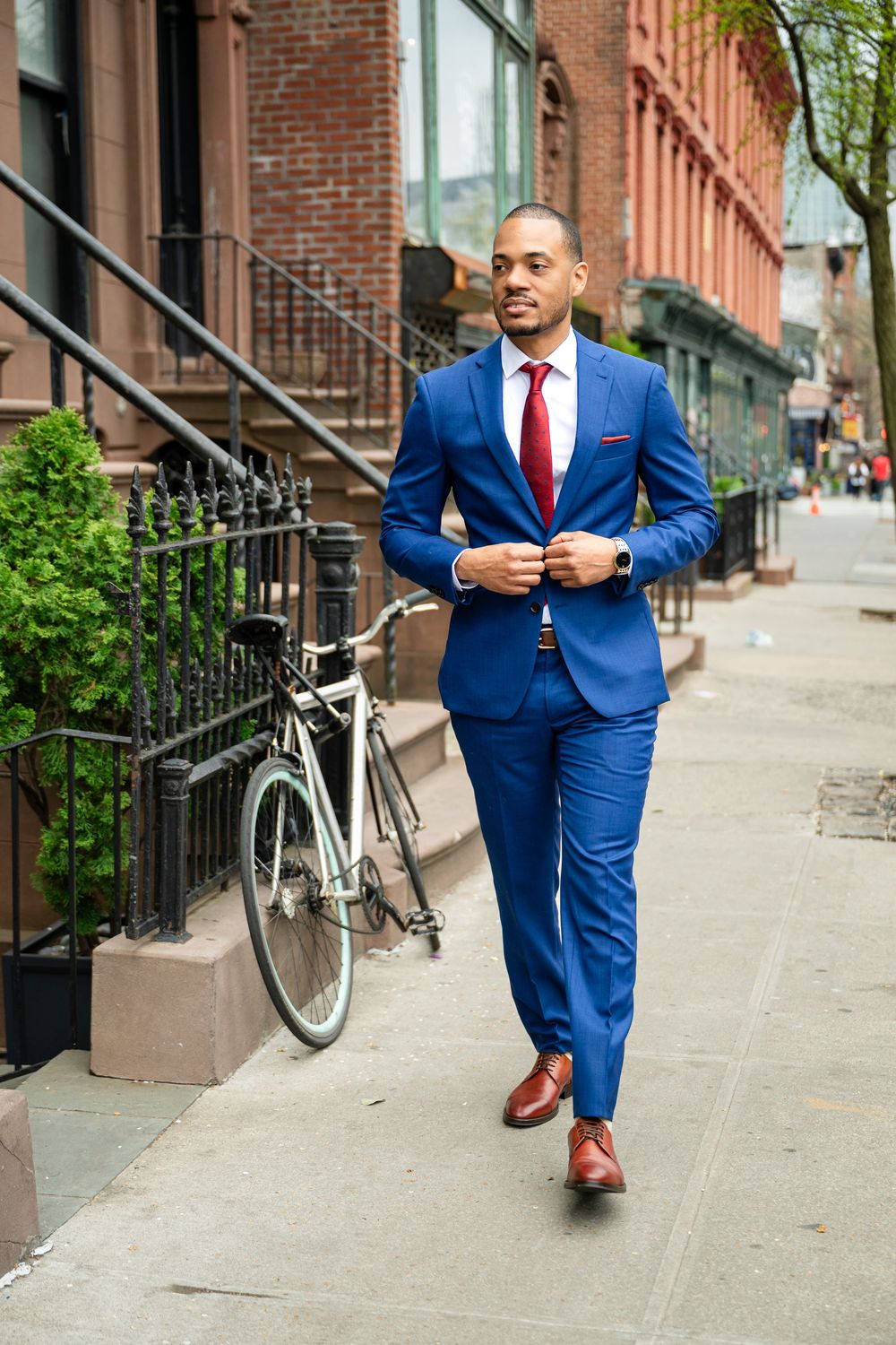 A person in a tailored royal blue suit and red tie walks confidently on a Brooklyn brownstone sidewalk next to a bicycle.