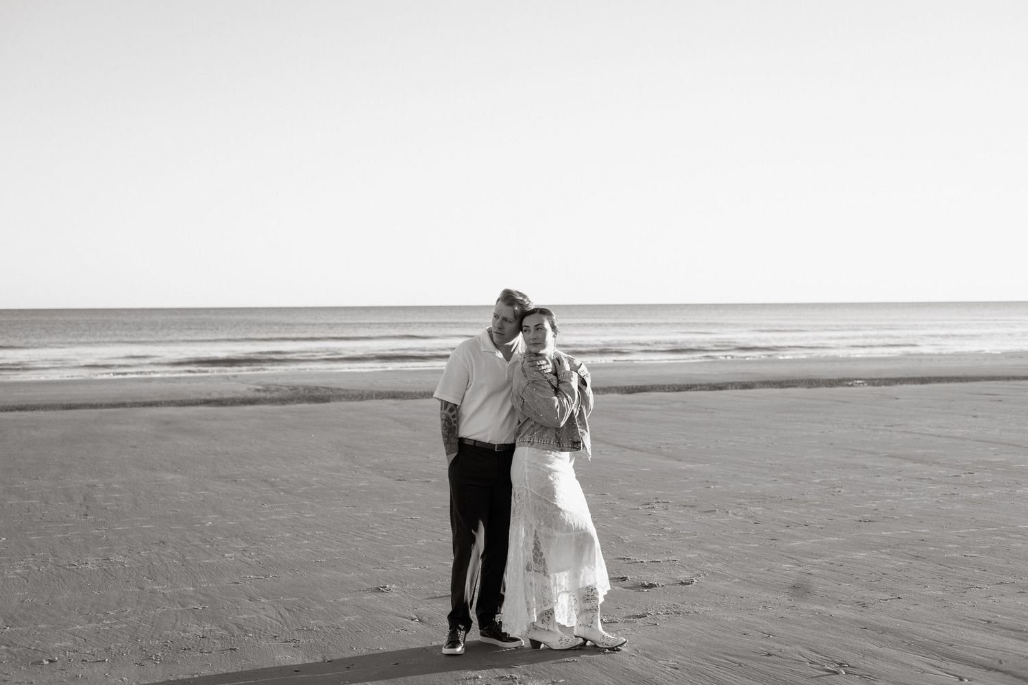 A romantic couple embraces while standing on a serene beach at sunset in black and white photography.
