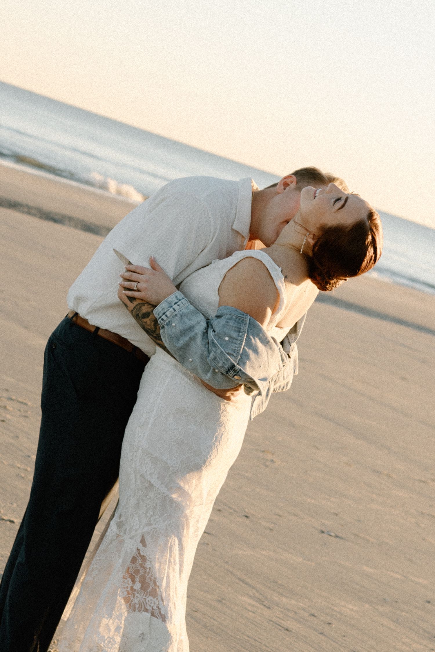 A couple in white and denim embrace romantically at sunset on a peaceful sandy beach.