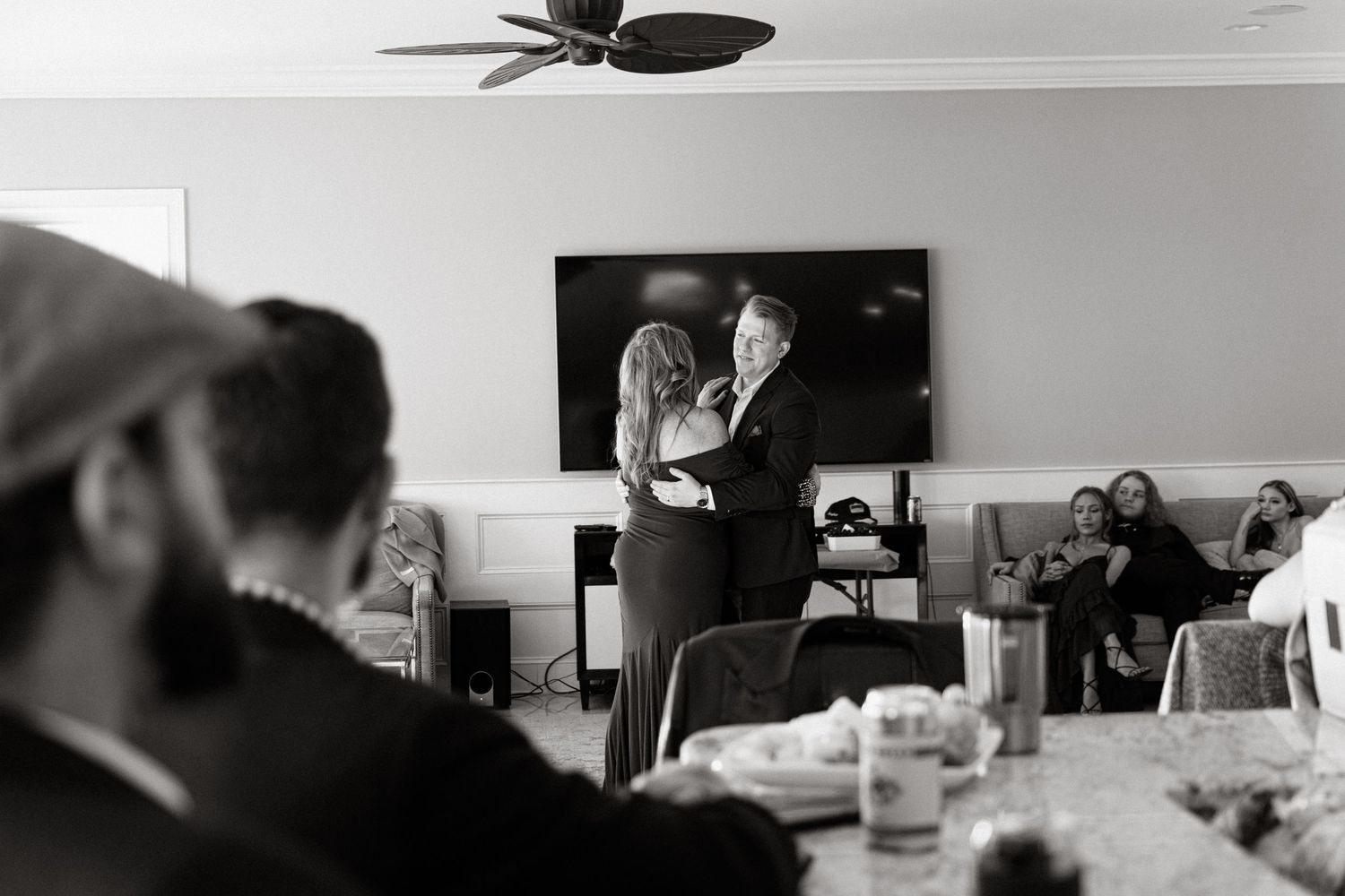 Black and white photo of people gathered in a dining room with a TV mounted on the wall during an event.
