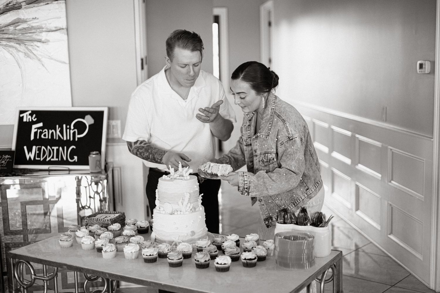 A couple cuts their wedding cake together while standing at a dessert table with cupcakes and a decorative sign.