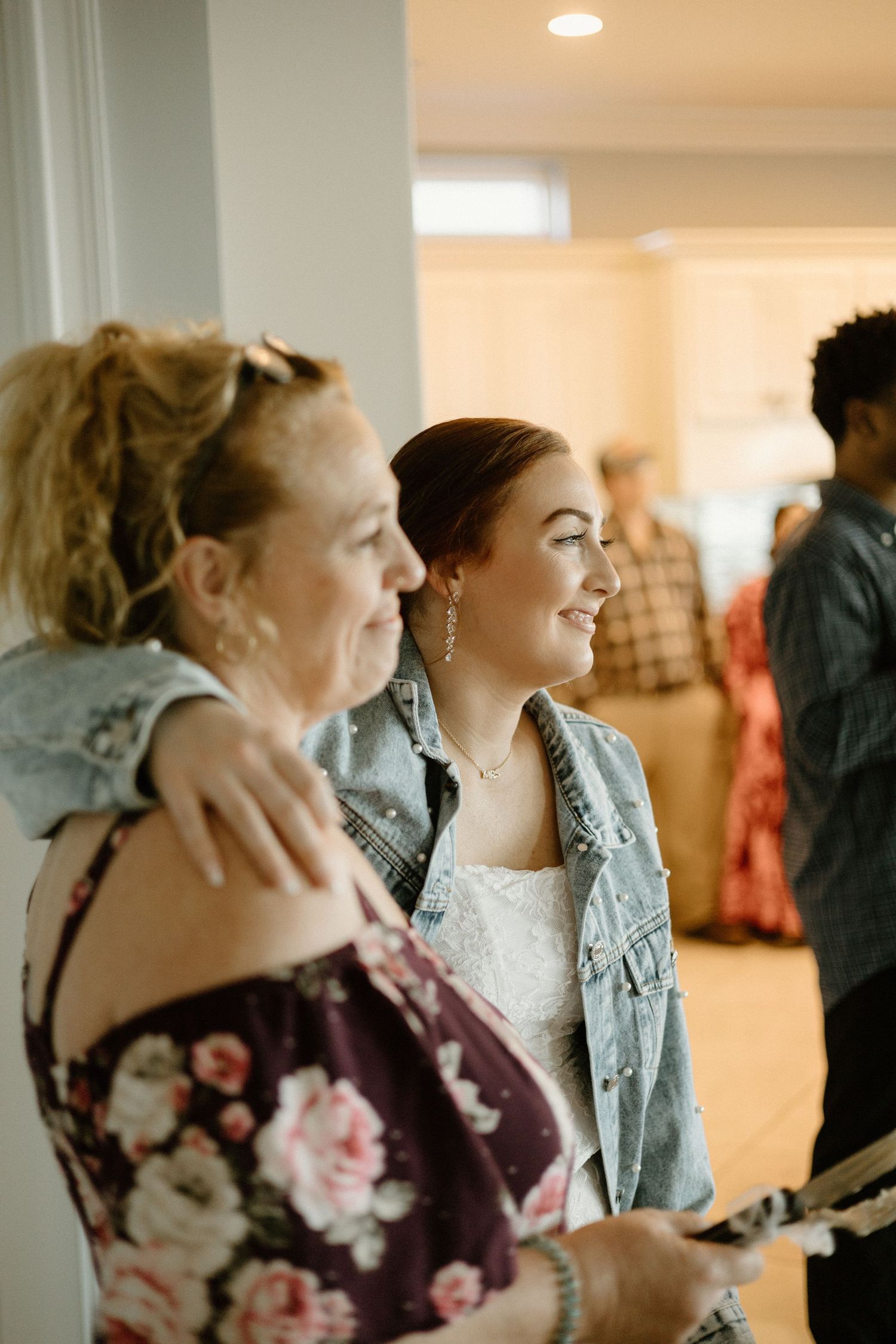 Two people share a joyful moment at an indoor gathering, one wearing a denim jacket and one in a floral print top.