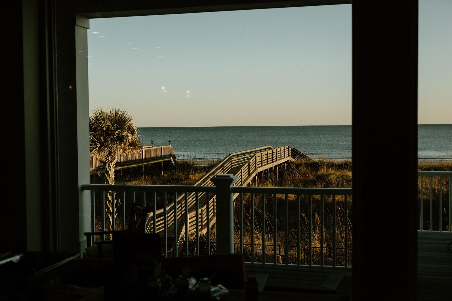 Wooden beach boardwalk leading to the ocean at sunset with palm trees and calm waters in view.