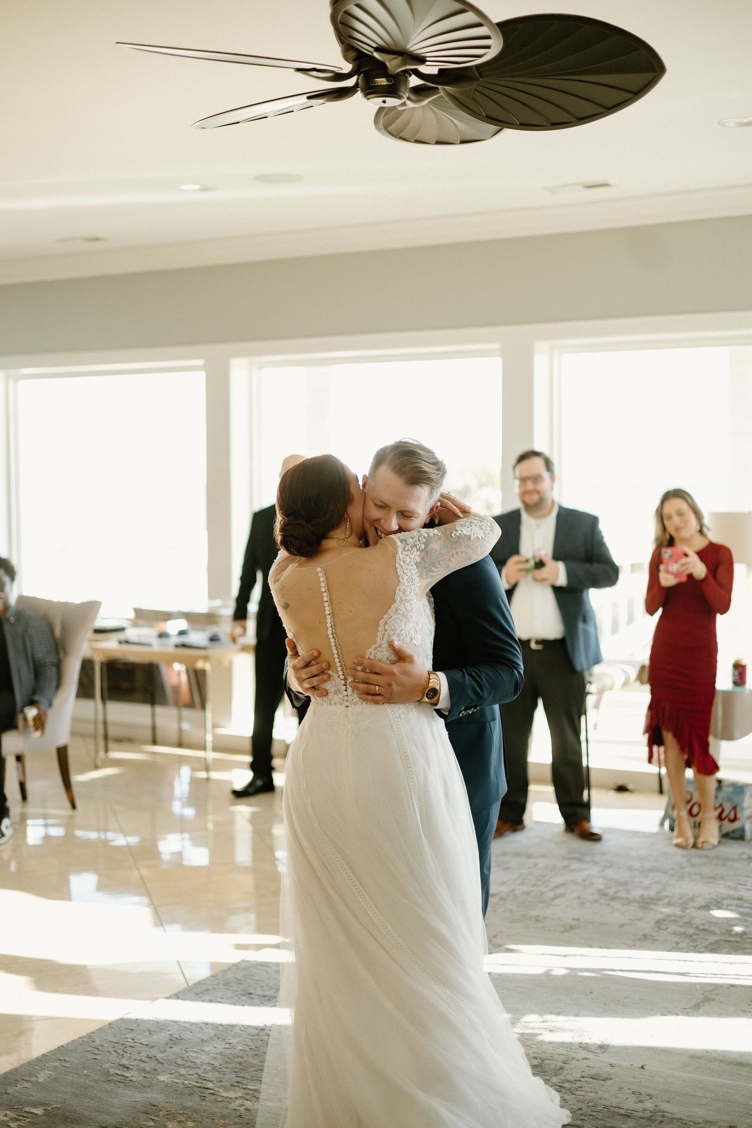 Wedding party guests line up to dance in sequence across a bright sunlit room with white floor-length windows.