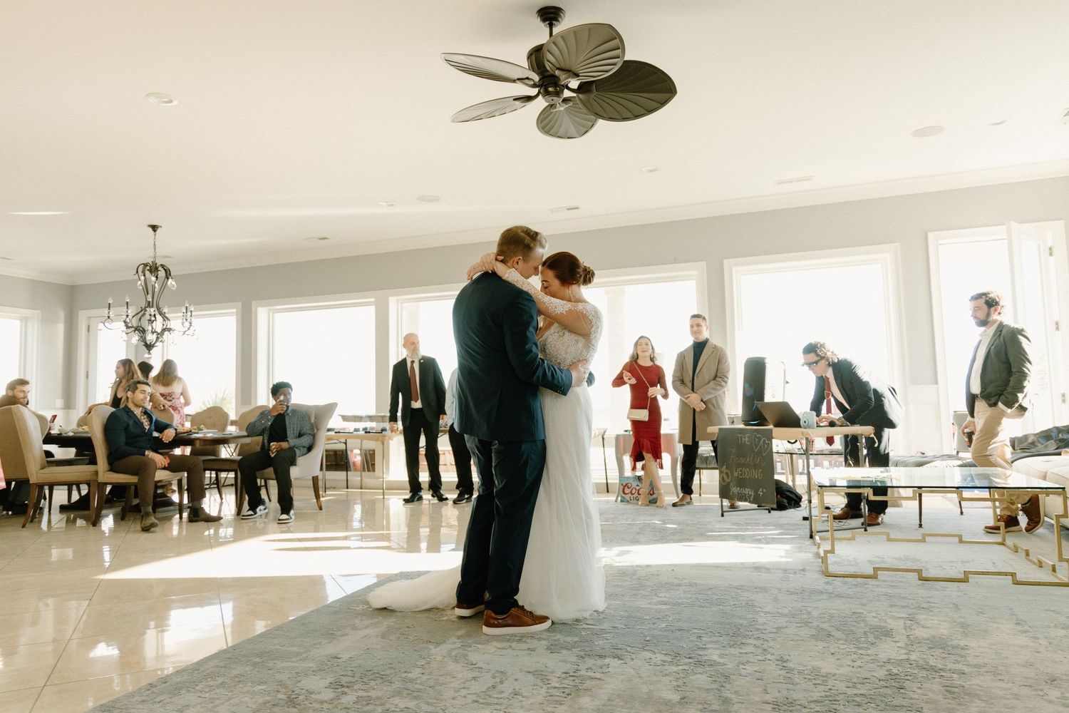 A couple shares a dance surrounded by guests in an elegant white reception venue with large windows.