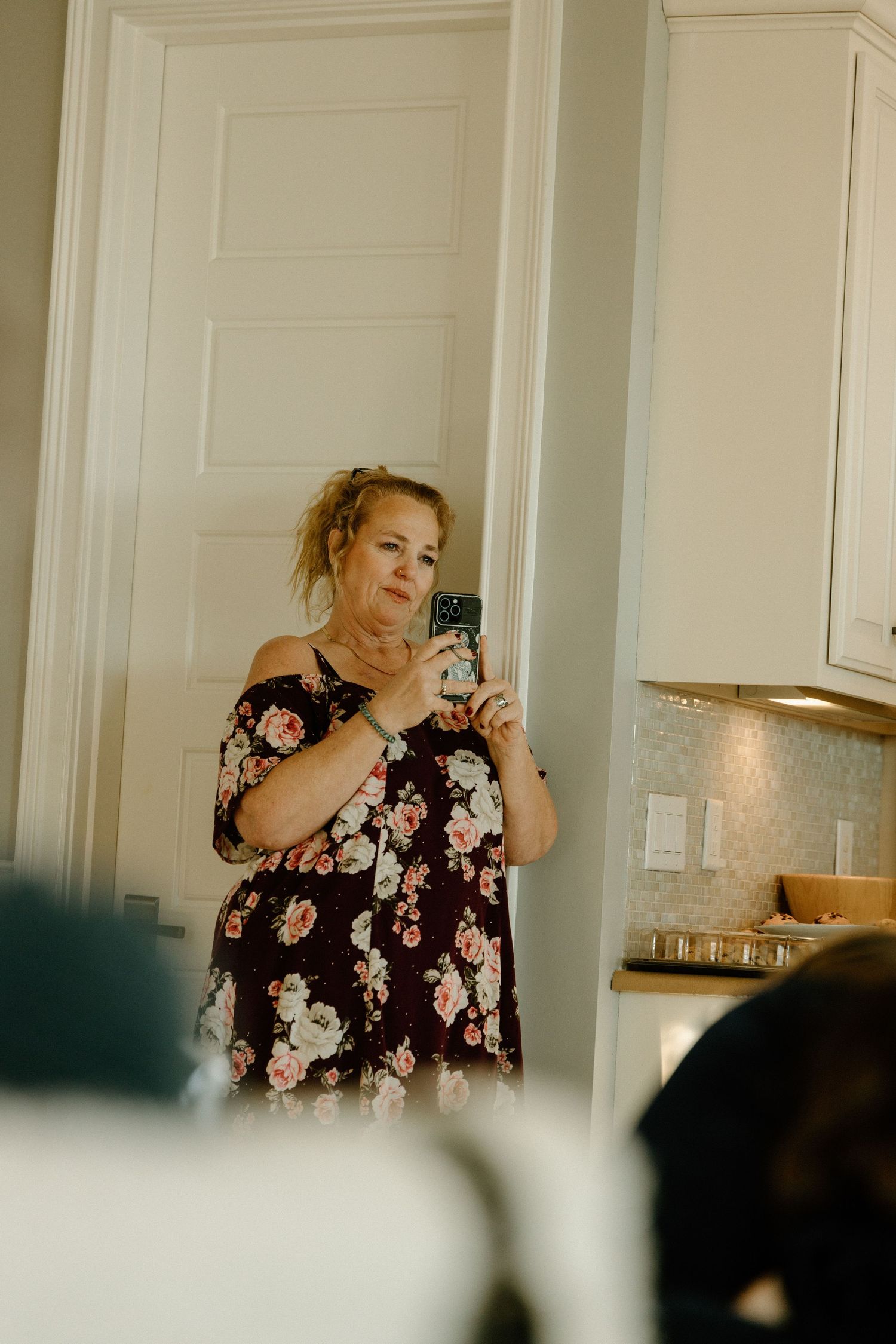 Person in a floral print dress takes a selfie in front of a light colored wall in a kitchen setting.