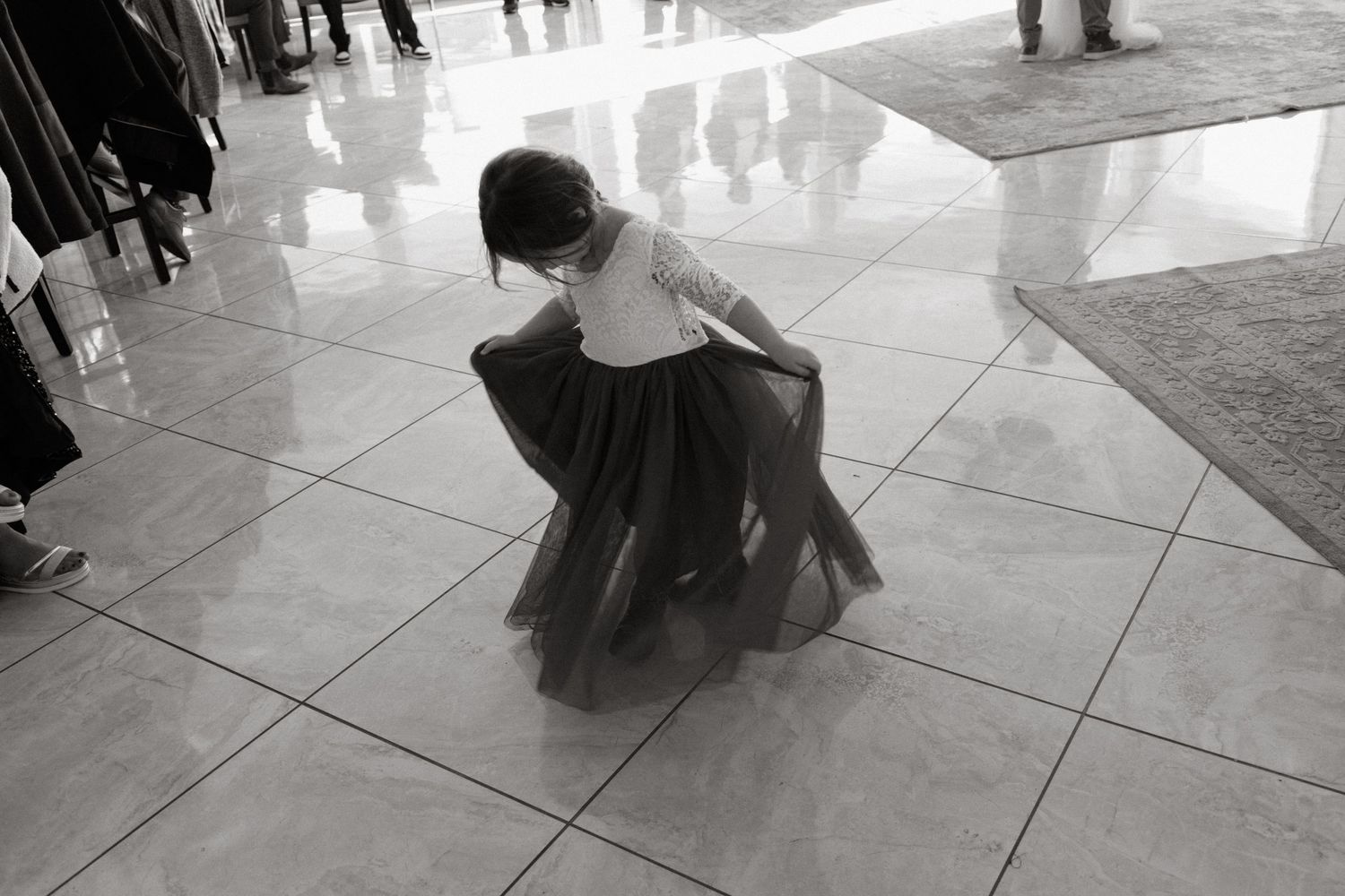 A dancer in a flowing dark dress crouches low on a reflective tiled floor in black and white photography.
