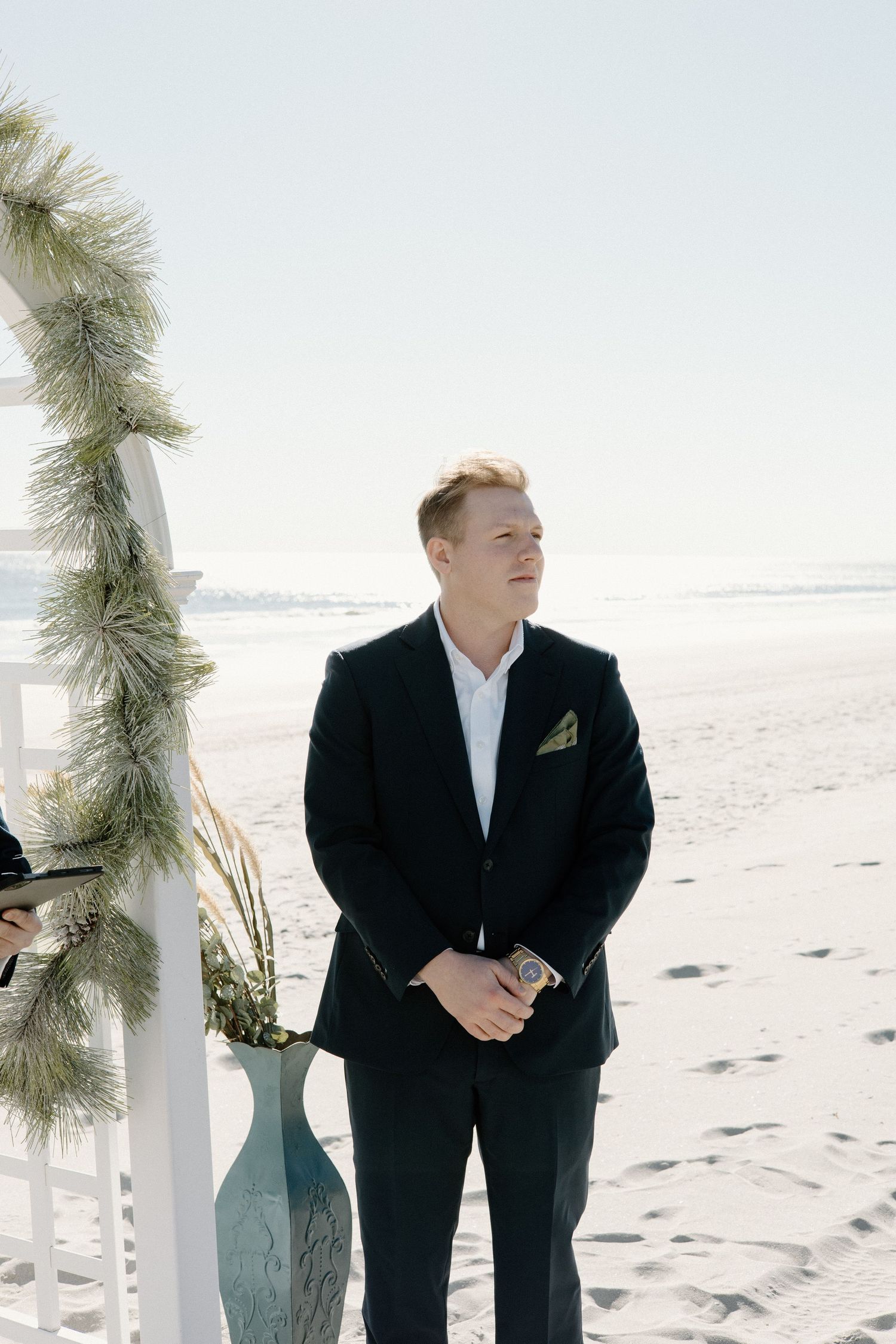 A groom in a black suit stands by a decorative arch with winter greenery on a sunny beach.