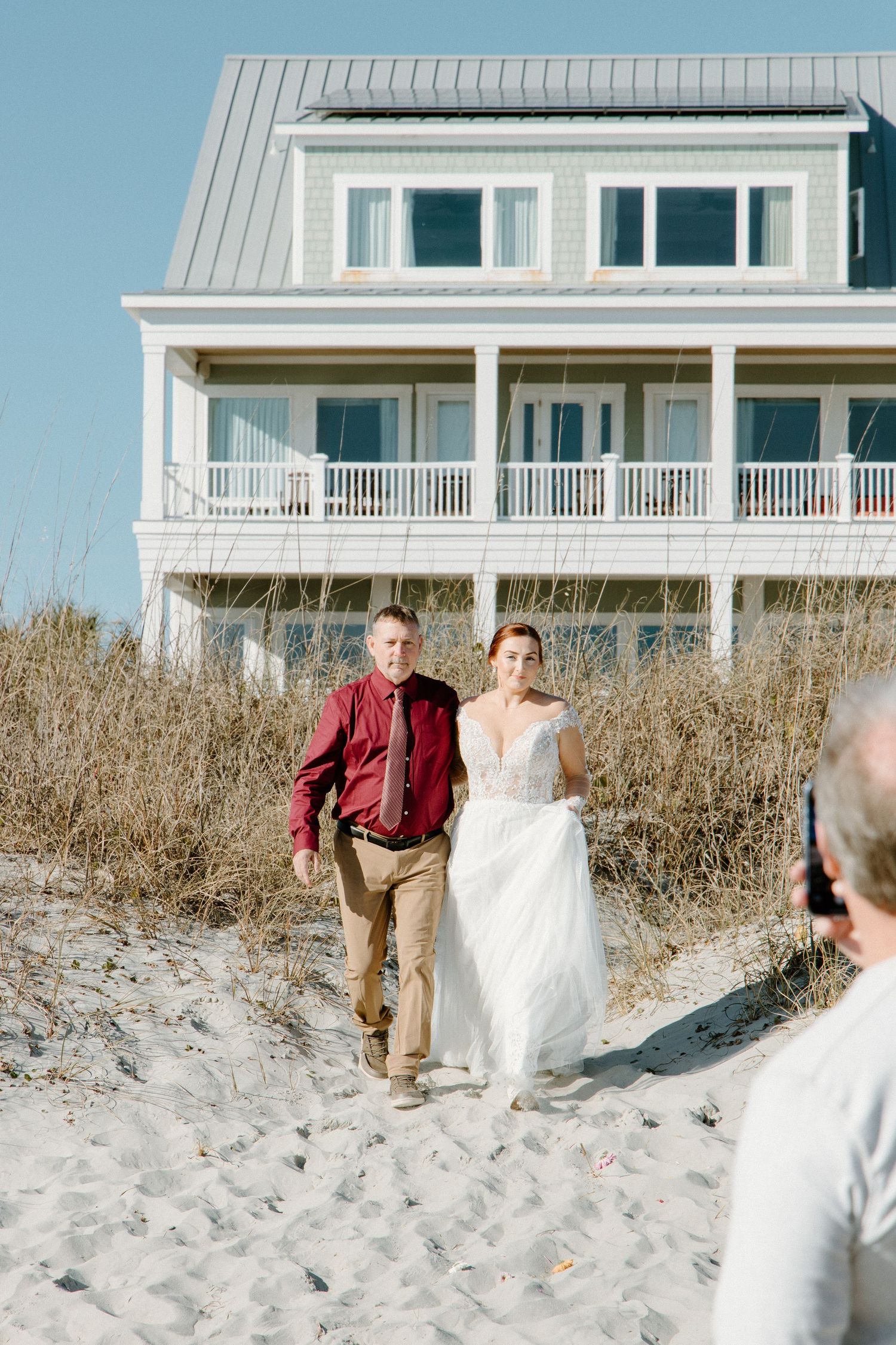 Beach house wedding scene with couple walking along sandy path near coastal home on sunny day.